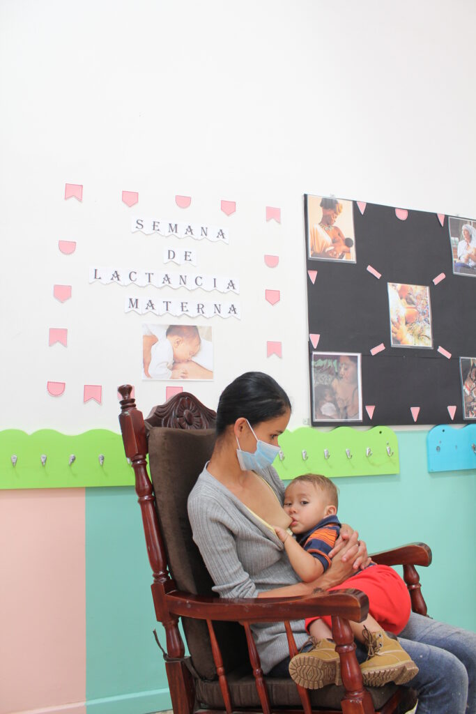 En la imagen se muestra una madre sentada en una silla amantando a su hijo